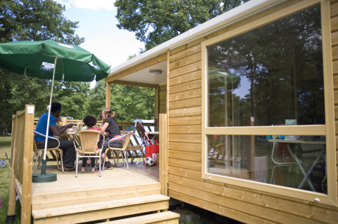 Family enjoying a meal on the deck of a wooden cabin at Huttopia Lac de Sillé, Glamping Pays de la Loire.