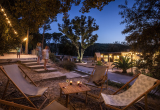 Outdoor glamping area at Huttopia Forêt de Janas with lounge chairs, lights, and guests at dusk.