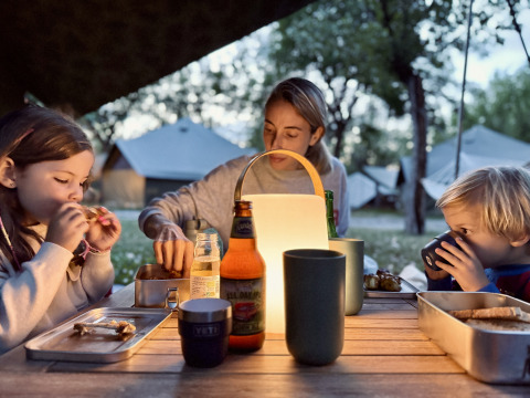 Familia cena al aire libre bajo una lámpara, acampando en Basecamps - Cabins Girona, con tiendas detrás.