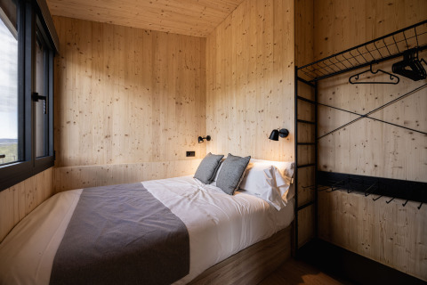 Basecamps - Cabins Girona glampingkamer met tweepersoonsbed, houten wanden en groot raam met uitzicht op natuur.