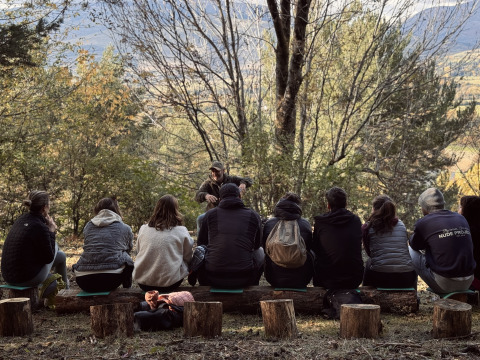 Een groep mensen zit op boomstammen in het bos bij Basecamps - Cabins Girona tijdens een buitenpresentatie.