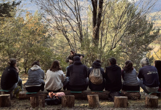 Un groupe de personnes assis sur des rondins en forêt à Basecamps - Cabins Girona écoute une présentation en plein air.