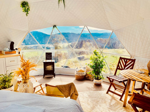 Interior de una cúpula glamping con grandes ventanas, muebles acogedores y vistas a campos y montañas.