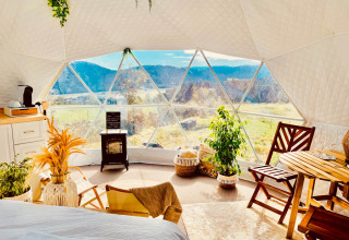 Interieur van een glamping dome met grote ramen, houten meubels en prachtig uitzicht op velden en bergen.
