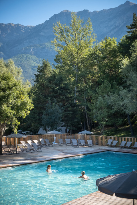 Piscina al aire libre con dos personas nadando, rodeada de árboles y montañas en Huttopia Lac de Serre-Ponçon.