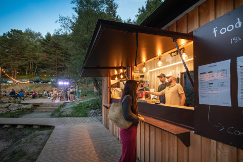 Vrouw bestelt eten bij buitenkraam op Camping Huttopia Lac de Serre-Ponçon, gezellige glamping sfeer.