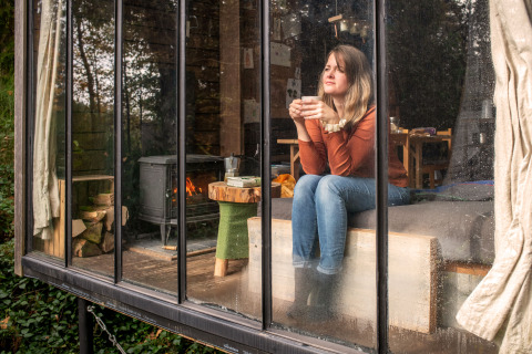 Mujer relajándose con té en un lodge Nutchel en las Ardenas, Bélgica, con paredes de vidrio y chimenea.