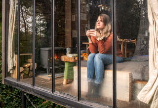 Femme détendue avec du thé dans une lodge Nutchel en Ardenne belge, murs vitrés et poêle visible.