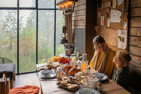 Madre e hijo desayunan en una acogedora cabaña con vistas al bosque en Nutchel Lodges, Ardenas, Bélgica.