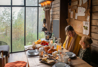 Madre e hijo desayunan en una acogedora cabaña con vistas al bosque en Nutchel Lodges, Ardenas, Bélgica.