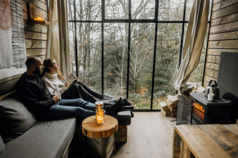 Couple relaxing in a Nutchel lodge in the Ardennes, Belgium, enjoying the forest view through large windows.