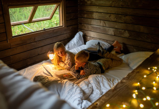 Two children reading a book together in a cozy wooden cabin with fairy lights, Nutchel Lodges Ardennes.