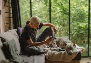Persoon ontspant op een bank met een hond bij Nutchel-lodges in de Ardennen, met glazen wand en groen uitzicht.
