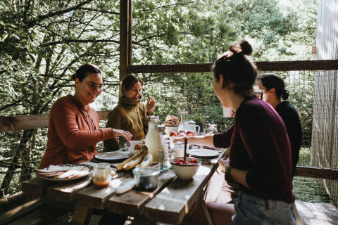 Vier Personen frühstücken gemeinsam an einem Holztisch im Grünen bei Nutchel - Lodges in den Ardennen, Belgien.