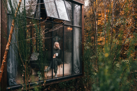 Donna con tazza seduta in una confortevole glamping lodge con grandi finestre a Nutchel, Ardenne, Belgio in autunno.