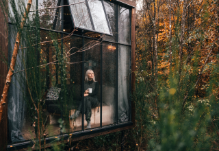 Donna con tazza seduta in una confortevole glamping lodge con grandi finestre a Nutchel, Ardenne, Belgio in autunno.