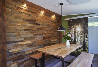 Espace repas glamping moderne avec mur en bois, table rustique, banc, lampe suspendue et bouquet de fleurs.