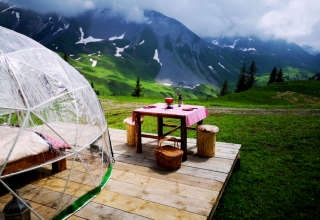 Glamping met glazen iglo, houten vlonder, picknicktafel en schilderachtig uitzicht op Alpenlandschap.