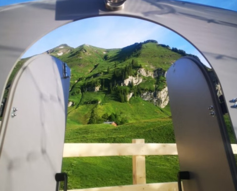 Vista desde Bubbelreis - Glazen Iglo Isenthal por puertas abiertas, mostrando montañas verdes y cielo azul.