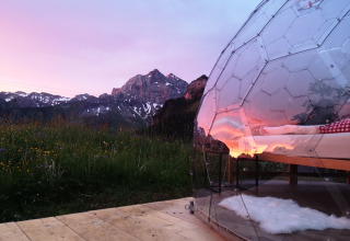 Alojamiento glamping de burbuja de vidrio en Isenthal, junto a montañas al atardecer con cielo rosado.