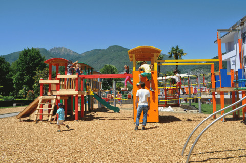 Des enfants jouent sur une aire de jeux colorée avec copeaux de bois, montagnes en arrière-plan au Campofelice Camping Village.