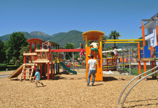 Des enfants jouent sur une aire de jeux colorée avec copeaux de bois, montagnes en arrière-plan au Campofelice Camping Village.