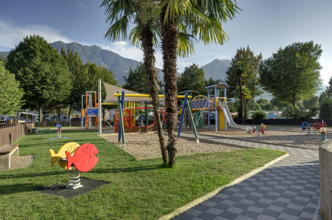 Parque infantil en Campofelice Camping Village - Igloo Tube Lago Maggiore con montañas y palmeras alrededor.