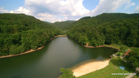 Vista del Campingpark Wiesenbeker Teich - Blokhutten Nedersaksen con lago, playa de arena y colinas boscosas.