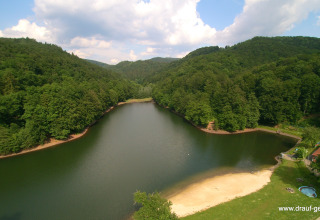 View of Campingpark Wiesenbeker Teich - Blokhutten Nedersaksen with a lake, sandy shore, and forested hills.