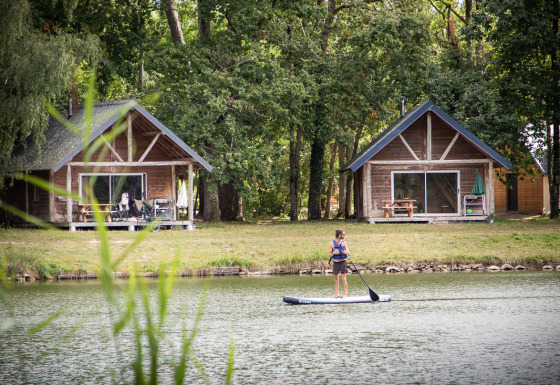 Zwei Holzhütten am See im Village Huttopia Lac de Rillé Glamping Val de Loire, Person auf einem SUP-Board.
