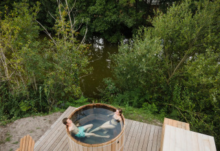 Two people relaxing in a hot tub on a wooden deck by a lake, surrounded by lush green trees.