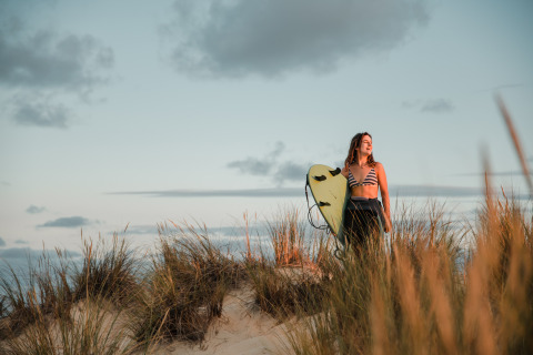 Giovane donna con tavola da surf tra le dune al tramonto, atmosfera glamping a Biscarrosse.
