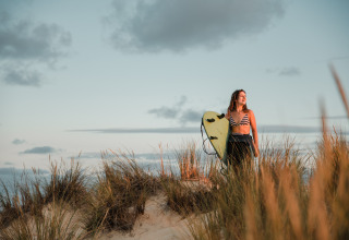 Junge Frau mit Surfbrett steht bei Sonnenuntergang in den Dünen, perfekte Stimmung in Biscarrosse.