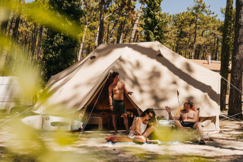 Three people relax in front of a glamping tent in Forestcamp Biscarrosse surrounded by trees and sunlight.