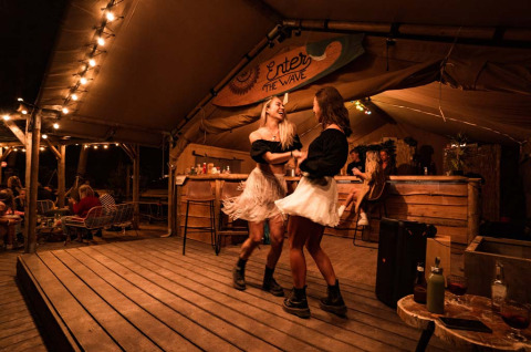 Twee vrouwen dansen op een verlicht houten podium in de gezellige glampingbar Enter the Wave – Forestcamp Biscarrosse.