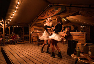 Zwei Frauen tanzen auf einer beleuchteten Holzterrasse in der gemütlichen Glampingbar Enter the Wave – Forestcamp Biscarrosse.