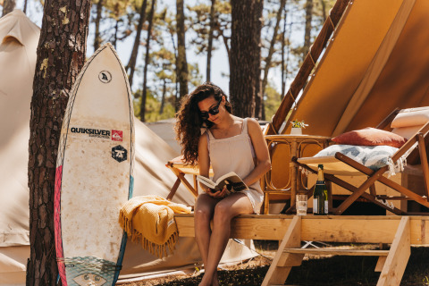 Vrouw leest een boek bij een glamping tent in het bos bij Forestcamp Biscarrosse met surfplank en wijn.