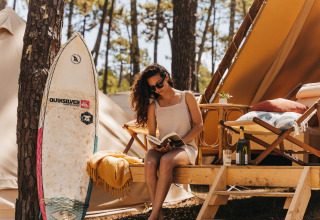 Kvinde læser bog ved glamping-telt i skoven hos Forestcamp Biscarrosse med surfbræt og vin.