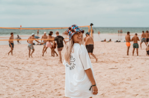 Woman in a white t-shirt and bucket hat smiles on the beach at Enter the wave – Forestcamp Biscarrosse glamping.