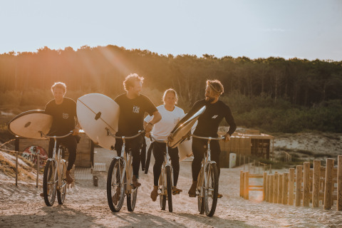 Vier mensen fietsen met surfplanken over een zandpad bij Enter the wave – Forestcamp Biscarrosse.