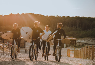 Vier personen fietsen met surfplanken op een zandweg bij Enter the wave – Forestcamp Biscarrosse.