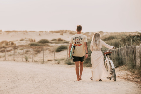 Par går barfodet på en sandsti ved skovcamping Forestcamp Biscarrosse med surfbræt og cykel.