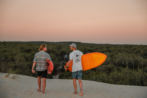 To surfere går med deres boards over sandklitter ved solnedgang nær Forestcamp Biscarrosse glamping.