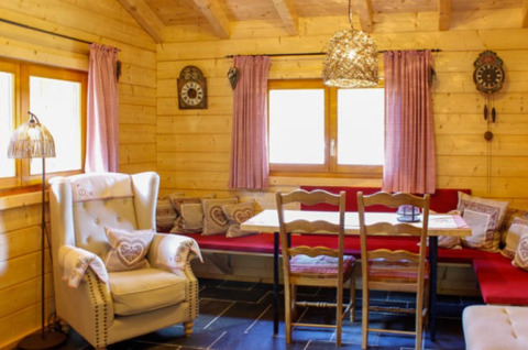 Acogedor interior de cabaña de madera con comedor, cojines y luz natural desde las ventanas.