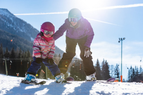 Un adulto y un niño esquían juntos en una pendiente nevada, rodeados de montañas, bosque y sol.