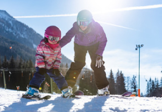 Un adulto e un bambino sciano insieme su una pista innevata con montagne e foresta al sole.