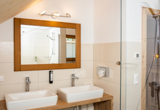 Baño moderno con lavabo doble, gran espejo y detalles en madera en HOCHoben - Chalets Oostenrijk glamping.