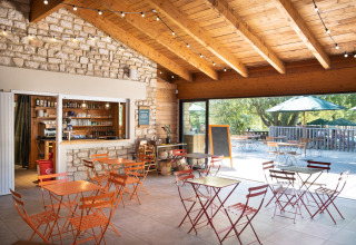 Acogedora cafetería con techo de madera y pared de piedra en Huttopia Le Moulin - Glamping Ardèche, mesas afuera.