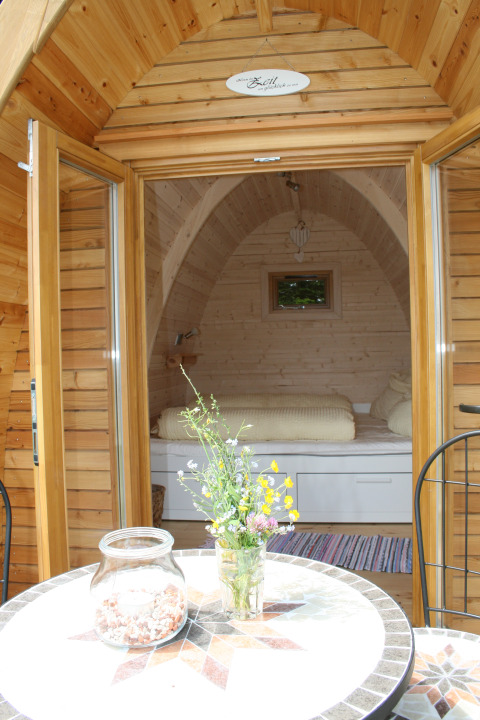 Ingresso di un’accogliente pod di legno con letto, tavolo e fiori, glamping Äckerhof nella Foresta Nera.