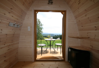 View from inside a wooden glamping pod with open door to a patio set and scenic outdoor landscape.